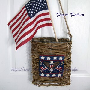 May include: A woven wicker basket with a blue and white needlepoint design featuring an eagle and the words "1776". The basket is hanging from a hook on a white door with a small American flag hanging next to it. The text "Paper Pattern" is visible in the image.