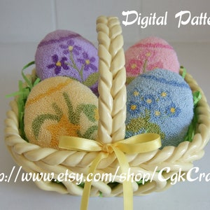 Peut inclure: Un panier en osier blanc avec un nœud en ruban jaune contient quatre œufs de Pâques brodés. Les œufs sont décorés de motifs floraux dans des tons de violet, rose, bleu et jaune.