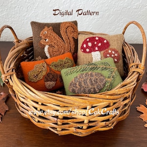 Pode incluir: Quatro pequenos travesseiros de feltro com bordados temáticos de outono são exibidos em uma cesta de vime. Os travesseiros apresentam um esquilo, um cogumelo, bolotas e folhas, e uma pinha. A cesta tem duas alças e está sentada em uma superfície de madeira.