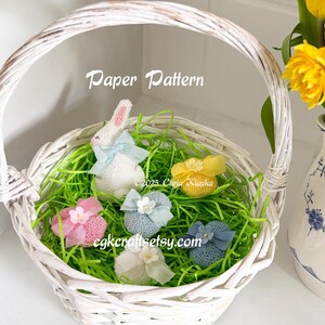 May include: A white wicker basket filled with green grass and pastel colored Easter eggs. The eggs are decorated with bows and flowers. A white bunny with a blue bow sits in the basket. The text "Paper Pattern" is written above the basket.