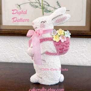 May include: A white bunny figurine with a pink basket filled with colorful flowers. The bunny has pink accents on its ears and cheeks, and a pink ribbon around its neck. The text "Digital Pattern" is visible.