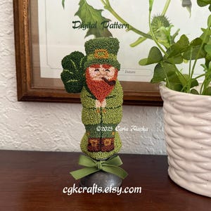 Puede incluir: Una figura de duende verde hecha a mano, con barba roja y pipa, sosteniendo un trébol. La figura está adornada con un lazo verde y se asienta sobre una base de metal. La imagen incluye el texto "Digital Pattern" y "cgkcrafts.etsy.com".