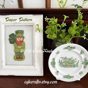 May include: White frame with the text "Paper Pattern" above a needlepoint image of a leprechaun holding a pot of gold. The image is on a white background.
