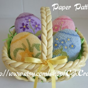 May include: A white wicker basket with a braided handle holds four fabric Easter eggs. The eggs are decorated with floral patterns in shades of purple, pink, blue, and yellow. The basket is filled with green grass.