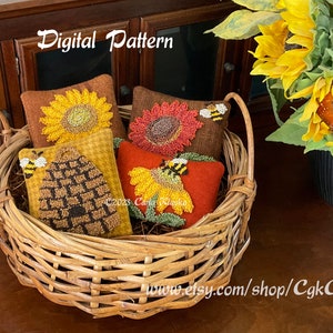 May include: A wicker basket filled with four small, hand-stitched felt pillows. The pillows feature a beehive, a sunflower, and two red and yellow sunflowers. The basket is on a wooden surface with a bouquet of sunflowers in a black vase.