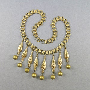 May include: A gold-tone necklace with a chain link design and multiple dangling charms. Each charm is a teardrop shape with a textured floral design and a round bead at the bottom.
