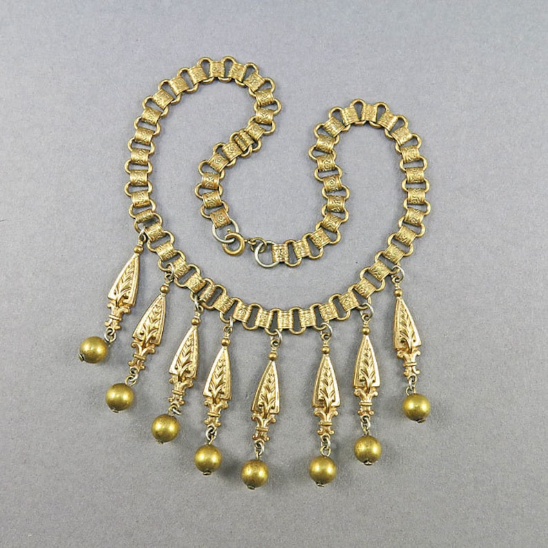 May include: A gold-tone necklace with a chain link design and multiple dangling charms. Each charm is a teardrop shape with a textured floral design and a round bead at the bottom.