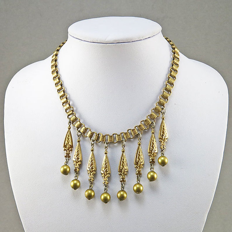 May include: A gold-tone necklace with a chain and multiple dangling teardrop-shaped charms. Each charm has a detailed floral design and is accented with a round gold bead.