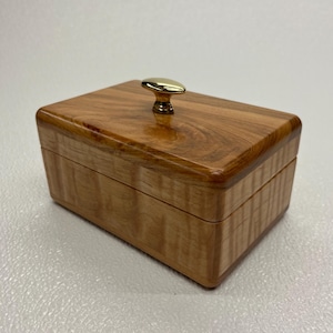 May include: A rectangular wooden box with a polished finish and a gold-colored knob on the lid. The box has a natural wood grain pattern and rounded corners, suggesting a handcrafted design.