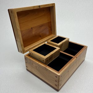 May include: A wooden storage box with a hinged lid, open to show three compartments and two removable boxes. The box is made of light and dark wood, with a dark interior. The box is likely used for storing items.