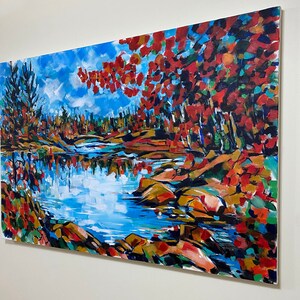 May include: A painting of a lake scene with red, orange, and yellow fall foliage on the banks. The water is blue and reflects the sky. There are rocks in the water and trees in the background.