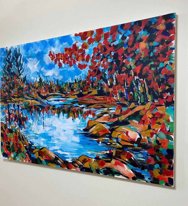 May include: A painting of a lake scene with red, orange, and yellow fall foliage on the banks. The water is blue and reflects the sky. There are rocks in the water and trees in the background.