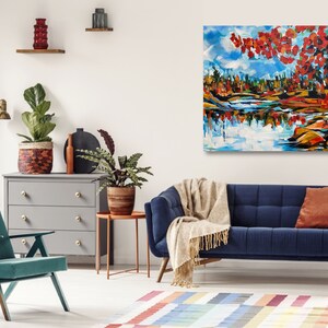 May include: A blue velvet sofa with a throw blanket and two pillows in a living room setting. A painting of a lake scene with red and orange trees hangs on the wall above the sofa.