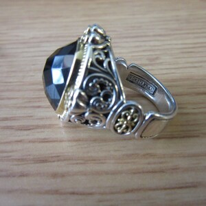 May include: A silver ring featuring a large black gemstone and intricate gold filigree detailing. The ring is inscribed with "Konstantino" on the inside band.