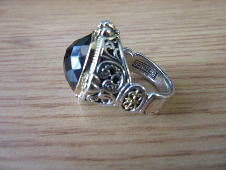 May include: A silver ring featuring a large black gemstone and intricate gold filigree detailing. The ring is inscribed with "Konstantino" on the inside band.
