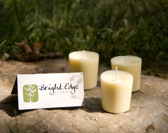 May include: Three ivory-colored pillar candles with white wicks are arranged on a stone surface. A small card with the text "Bright Edge Candles" and a tree logo is in front of the candles. The background is blurred greenery.