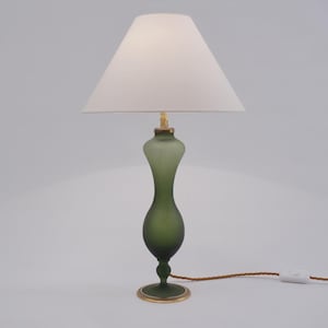May include: A green glass table lamp with a white lampshade. The lamp has a slender, hourglass-shaped body and a gold-colored base. The cord is brown with a white switch. The lamp is approximately 20 inches tall.