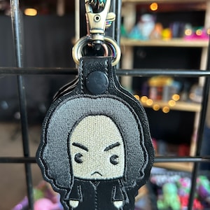 May include: Black embroidered keychain featuring a cartoon character with a serious expression. The character is wearing a black robe and has black hair.