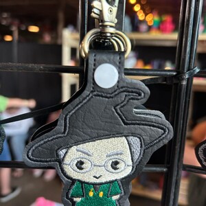 May include: Black leather keychain with an embroidered cartoon character wearing a black hat, glasses, and a green shirt.