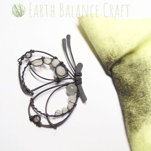 K&ouml;nnte beinhalten: Eine Draht-Schmetterlings-Skulptur mit wei&szlig;en Perlen und einer hellbraunen Oberfl&auml;che. Der Schmetterling befindet sich auf einer wei&szlig;en Oberfl&auml;che neben einem St&uuml;ck gelbem Filz.