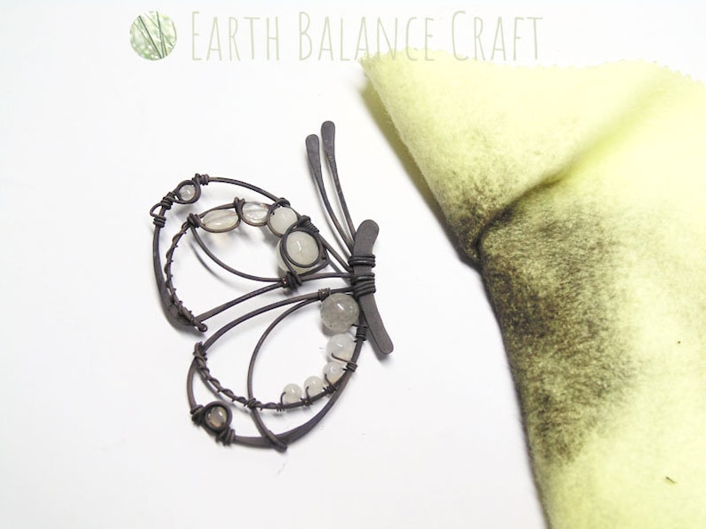 K&ouml;nnte beinhalten: Eine Draht-Schmetterlings-Skulptur mit wei&szlig;en Perlen und einer hellbraunen Oberfl&auml;che. Der Schmetterling befindet sich auf einer wei&szlig;en Oberfl&auml;che neben einem St&uuml;ck gelbem Filz.