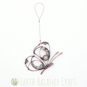 K&ouml;nnte beinhalten: Ein Schmetterlings-Anh&auml;nger aus Kupferdraht mit wei&szlig;en Edelstein-Akzenten. Der Schmetterling h&auml;ngt an einem d&uuml;nnen Kupferdraht.