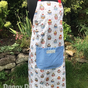 May include: A white apron with a red and white cupcake pattern. The apron has a blue pocket with white polka dots and a white label that says "Dappy Doo Dah".