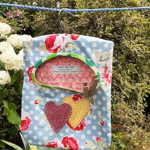Puede incluir: Una bolsa de lavandería azul y blanca con lunares, con estampado floral y dos corazones de tela. La bolsa está colgada de un tendedero y tiene una etiqueta que dice "Dappy Doo Dah By Sue Elsbury Made in Somerset, England."