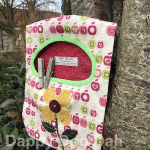 Puede incluir: Un organizador colgante de tela blanca con estampado de manzanas verdes y rosas. El organizador tiene un bolsillo rojo con una etiqueta que dice "DAPPY DOO DAH by Sue Elbury Made in Somerset, England".