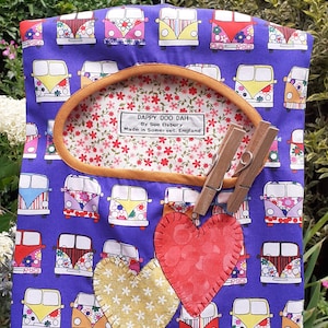 Puede incluir: Un organizador colgante de tela morado con un patrón de furgonetas Volkswagen vintage de colores. El organizador tiene dos bolsillos en la parte delantera, uno con un corazón rojo y otro con un corazón amarillo. El organizador está colgado de una percha de metal y tiene una etiqueta que dice "Dappy Doo Dah by Sue Elsbury Made in Somerset, England."