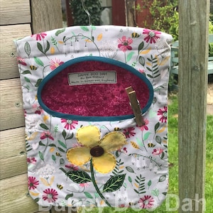 Puede incluir: Una bolsa de lavandería de tela blanca con estampado floral y una aplicación de flor amarilla. La bolsa tiene un ribete azul turquesa y un forro rosa. La bolsa está colgada de una percha de metal y tiene una etiqueta que dice "Dappy Doo Dah by Sue Elsbury Made in Somerset, England".