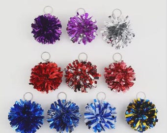Puede incluir: Una colección de pompones coloridos con fijaciones de anillo de metal plateado. Los pompones son de varios colores, incluyendo morado, rojo, azul, plateado y amarillo. Estos artículos decorativos se utilizan probablemente para animadoras u otras ocasiones festivas.