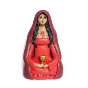 May include: A handcrafted figurine of a robed figure in red, holding a golden chalice. The figure has long black hair, green eyes, and a rose emblem on the chest. The text "L'Artefatto Esoterico" is at the bottom.