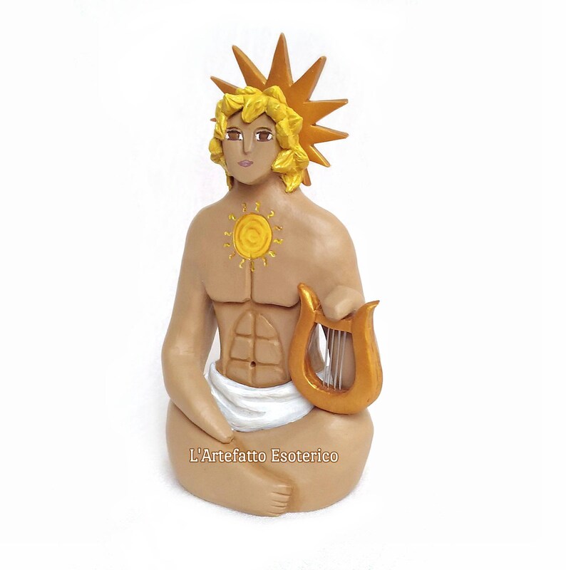 Puede incluir: Una figurita artesanal de una figura sentada con una corona solar, un s&iacute;mbolo solar en el pecho y sosteniendo una lira dorada. La figura est&aacute; hecha de arcilla y pintada en tonos marrones, dorados y amarillos. La figurita est&aacute; etiquetada como "L'Artefatto Esoterico".
