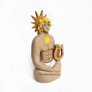 Puede incluir: Una figurita de cer&aacute;mica pintada a mano de una figura sentada con un halo solar, sosteniendo una lira. La figura tiene un torso musculoso y lleva una tela blanca. La figurita est&aacute; pintada en tonos marrones, dorados y amarillos.