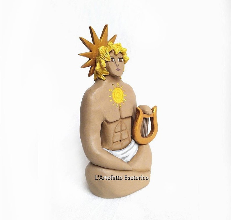 Puede incluir: Una figurita de cer&aacute;mica pintada a mano de una figura sentada con un halo solar, sosteniendo una lira. La figura tiene un torso musculoso y lleva una tela blanca. La figurita est&aacute; pintada en tonos marrones, dorados y amarillos.