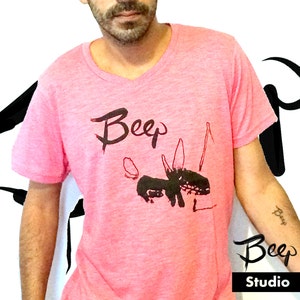 May include: Pink V-neck t-shirt with a black graphic design and the word "Beep" in a handwritten style. The design includes a stylized animal figure. The word "Beep" is also on the arm. The word "Studio" is below the word "Beep".