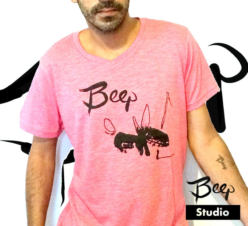 May include: Pink V-neck t-shirt with a black graphic design and the word "Beep" in a handwritten style. The design includes a stylized animal figure. The word "Beep" is also on the arm. The word "Studio" is below the word "Beep".