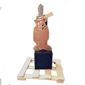 May include: A brown cartoon character cutout wearing a hat and holding a sign, mounted on a black cube. A feather is attached to the hat. The figure rests on a small wooden pallet.
