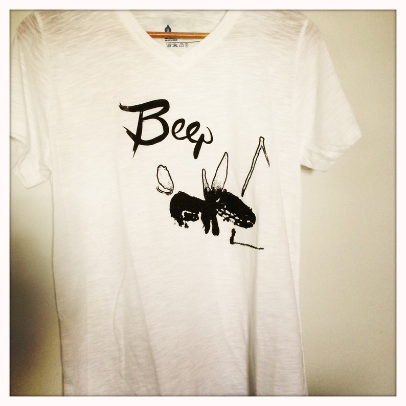 May include: White V-neck t-shirt with a black graphic of an animal and the word "Beep" in a stylized font. The shirt is hanging on a wooden hanger against a plain white wall.