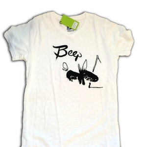 May include: Off-white t-shirt with a black graphic of an insect and the word "Beep" in a handwritten style. The shirt has short sleeves and a crew neck. A green tag is attached near the neckline.