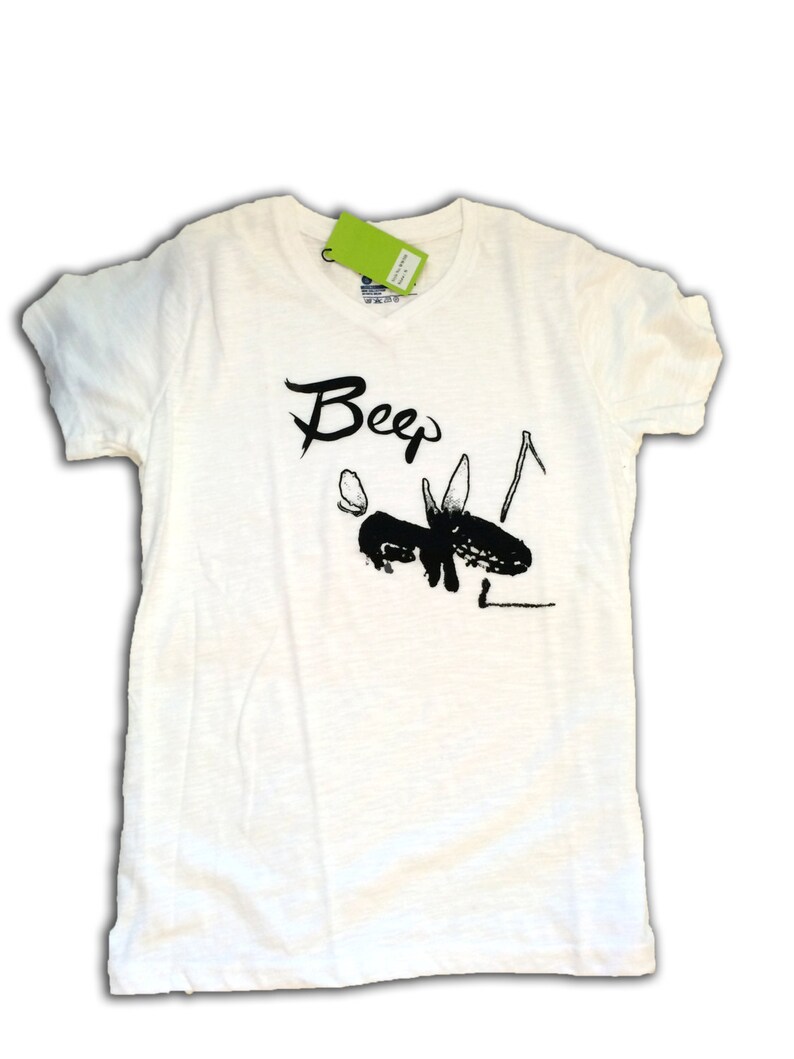 May include: Off-white t-shirt with a black graphic of an insect and the word "Beep" in a handwritten style. The shirt has short sleeves and a crew neck. A green tag is attached near the neckline.