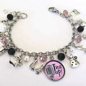 May include: A silver charm bracelet with a variety of charms, including a microphone with the text "Tits Up!" on a pink background, a telephone, a purse, a shoe, and a dress. The bracelet also features black and white beads.