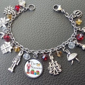 May include: Silver charm bracelet with various charms, including a snowflake, candy cane, gift box, and a charm that says "The Nutcracker". The bracelet also features red, yellow, and clear beads, and a ballerina charm. The text "IT'S GEEKtastic!" is in the upper right corner.
