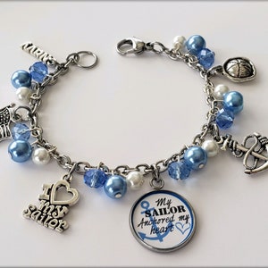 Puede incluir: Un brazalete de plata con abalorios azules y blancos y encantos, incluyendo un ancla, un coraz&oacute;n, una bandera y un encanto redondo con el texto "My Sailor Anchored my Heart".