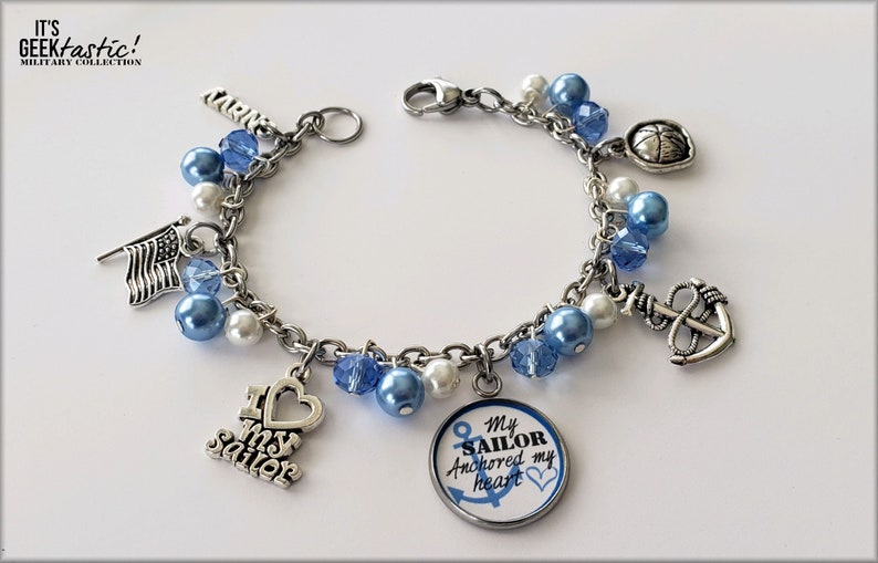 Puede incluir: Un brazalete de plata con abalorios azules y blancos y encantos, incluyendo un ancla, un coraz&oacute;n, una bandera y un encanto redondo con el texto "My Sailor Anchored my Heart".