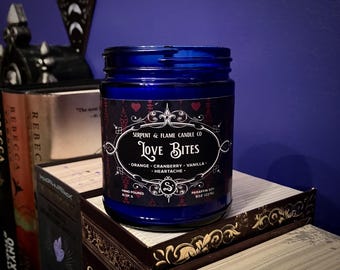 May include: A dark blue glass jar candle labeled "Love Bites" with the Serpent & Flame Candle Co. logo. The label lists scents: orange, cranberry, vanilla, and heartache. The candle sits on a stack of books with decorative edges.
