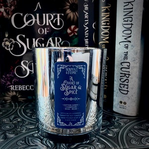 Puede incluir: Un portavelas de cristal transparente con un acabado plateado reflectante. La vela tiene una etiqueta azul que dice "A Court of Sugar & Spice" con notas de aroma. Libros en el fondo.