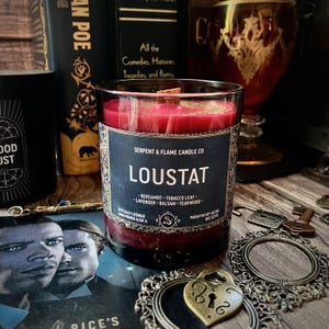 May include: A glass candle with a dark red wax and a wooden wick. The candle has a black label with the name "LOUSTAT" and the words "SERPENT & FLAME CANDLE CO." The scent notes are listed as bergamot, tobacco leaf, lavender, balsam, and teakwood.