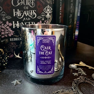 May include: A silver-colored candle in a glass jar with a purple label that reads "Serpent & Flame Caer aka The Cat." The candle is 11.5 oz (326g) and has a scent of violet, night air, woods, citrus, spearmint, and musk. The candle is next to books.